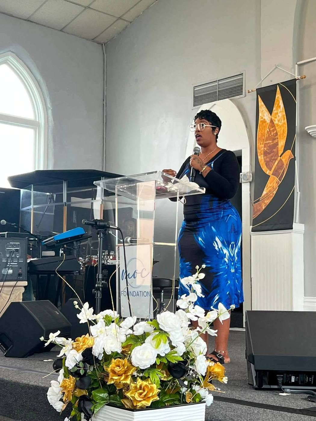 Speaker in a black top and blue skirt at the podium beside the golden dove banner.