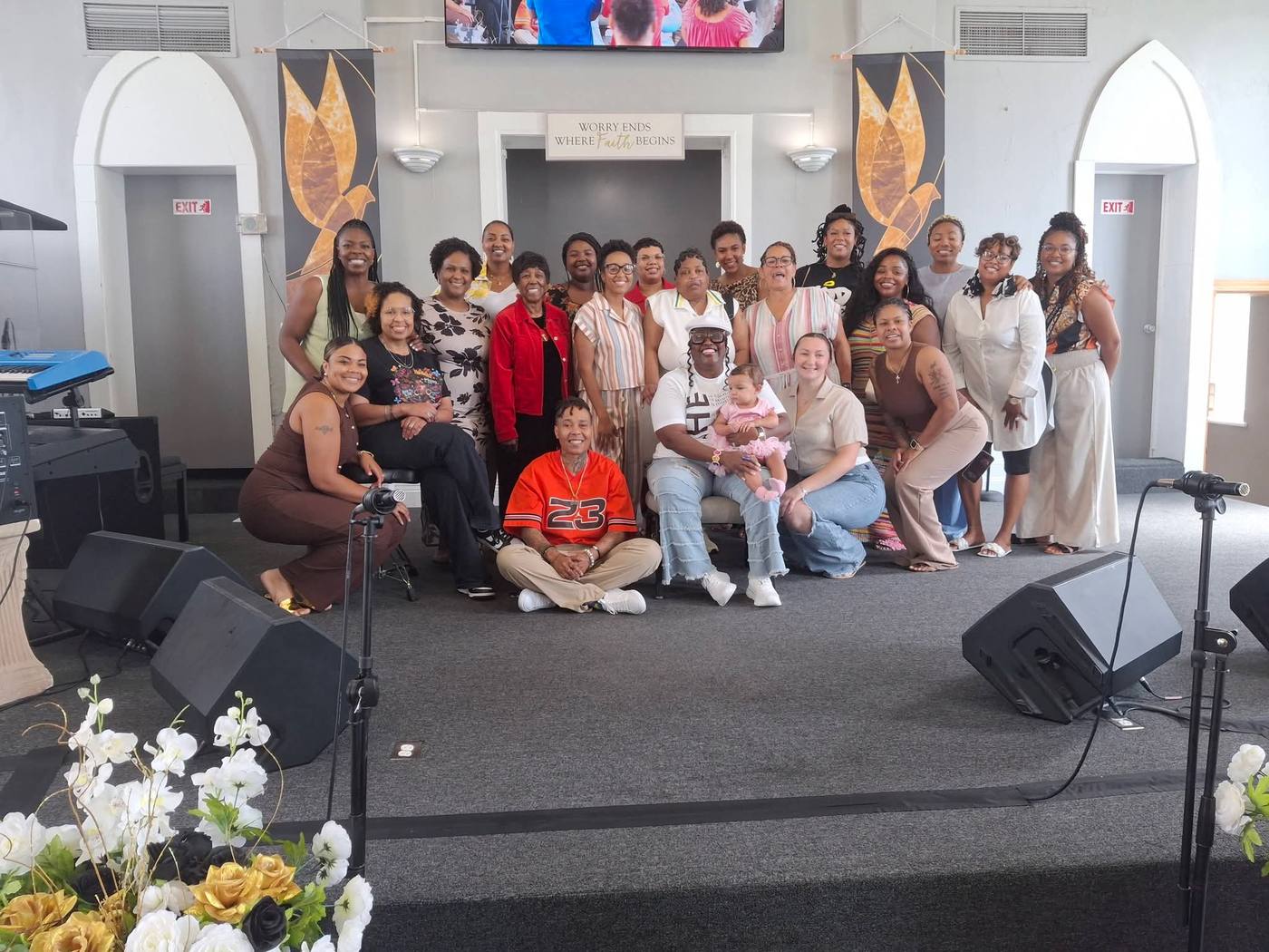 The full group of attendees gathered on stage at the close of the Breaking Bread & Chains Experience, beneath a sign reading 'Worry Ends Where Faith Begins.'