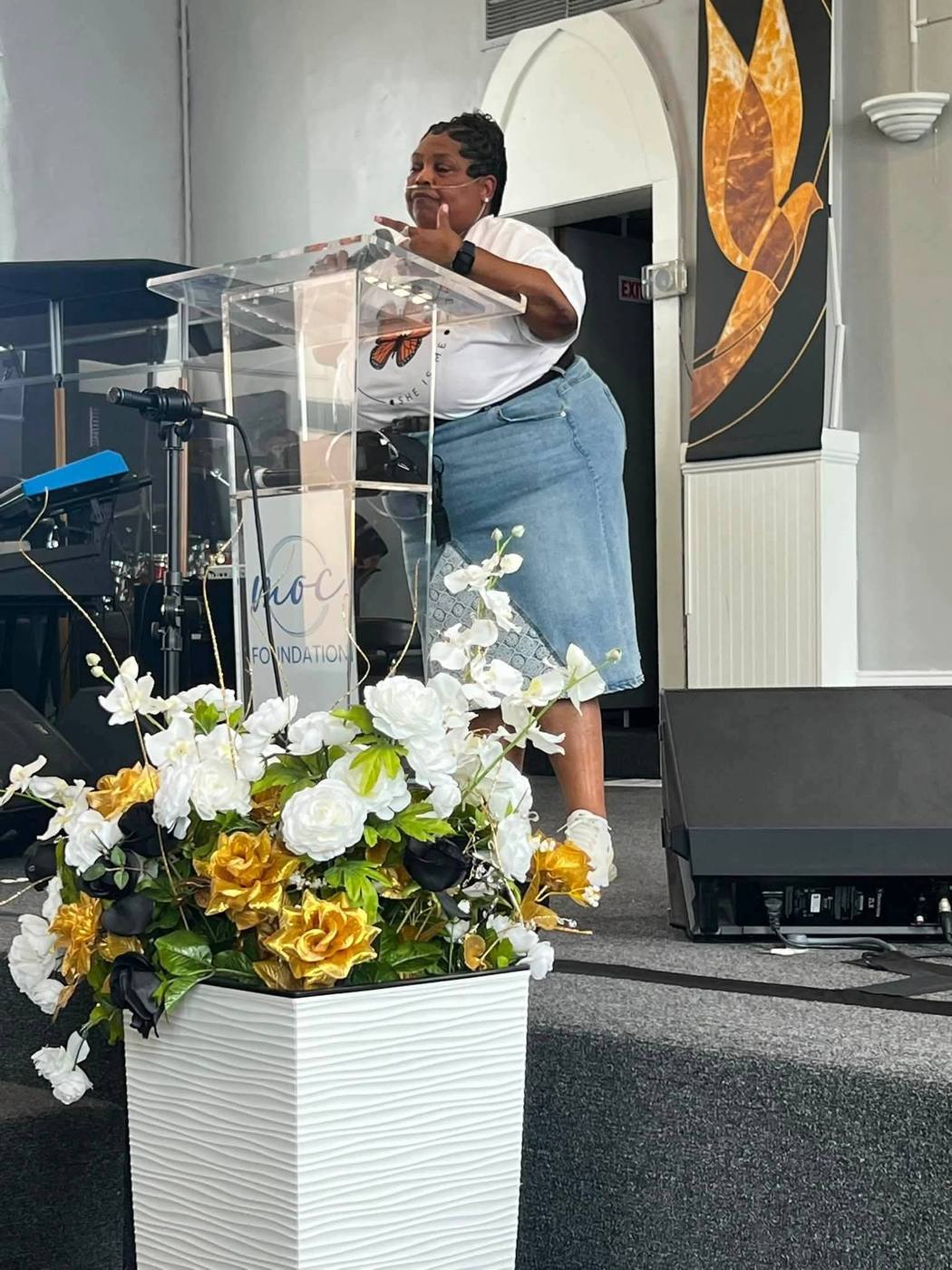 Speaker in a She Is Me butterfly tee gesturing while speaking at the podium.