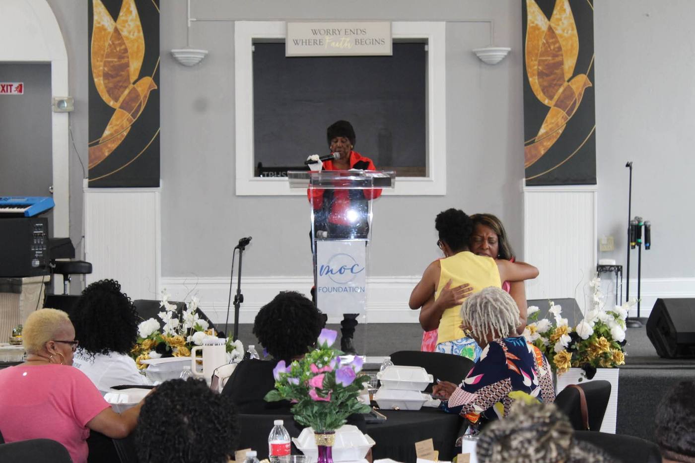 Speaker at the podium as two women embrace nearby in an emotional moment.