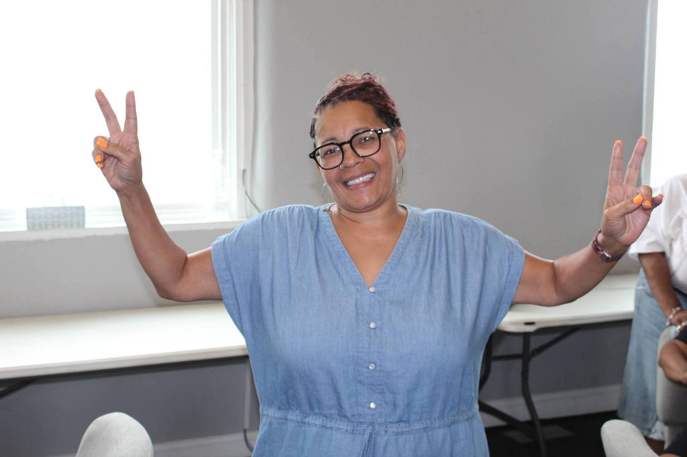 A woman in a chambray dress smiling with both hands raised in peace signs.